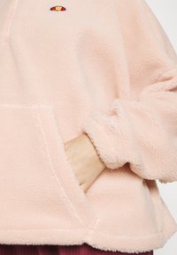 Main dans la poche d’une veste polaire rose clair avec des poignets côtelés et un petit logo brodé rouge et jaune près du col.