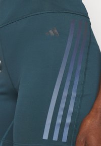 Gros plan sur un legging de sport bleu sarcelle foncé avec trois bandes sombres et un petit logo Adidas sur le haut de la cuisse, porté par une personne à la peau foncée.