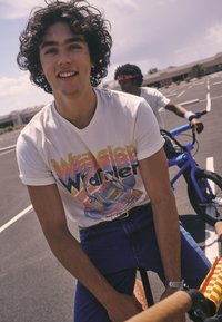 White t-shirt with colorful graphic design featuring a vintage car and headphones, paired with blue jeans and a silver watch.