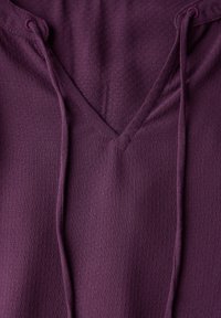 Haut à capuche violet en V, présentant un tissu texturé avec de subtiles perforations et des cordons, mettant en avant un design décontracté et fonctionnel.