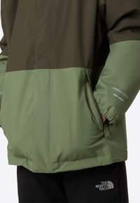 Jachetă impermeabilă verde măsliniu și verde deschis, cu manșete elastice, buzunare laterale și detaliu reflectorizant cu logo-ul „DRYVENT”.