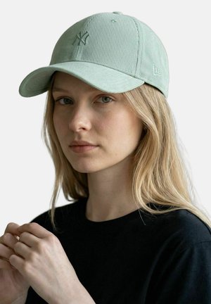 Junge Frau mit langen blonden Haaren trägt eine hellgrüne Cord-Baseballkappe und ein schwarzes Hemd, Hände nahe der Brust gefaltet.