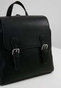 Mochila de cuero negra con un diseño estructurado, que cuenta con herrajes plateados, doble hebilla y un asa en la parte superior para llevarla.