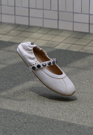 Fehér bőrből készült lapos cipő rugalmas hátsó résszel és pánttal, amelyet nagy fekete gombok díszítenek, szürke aszfalton, csempézett fal háttérrel.