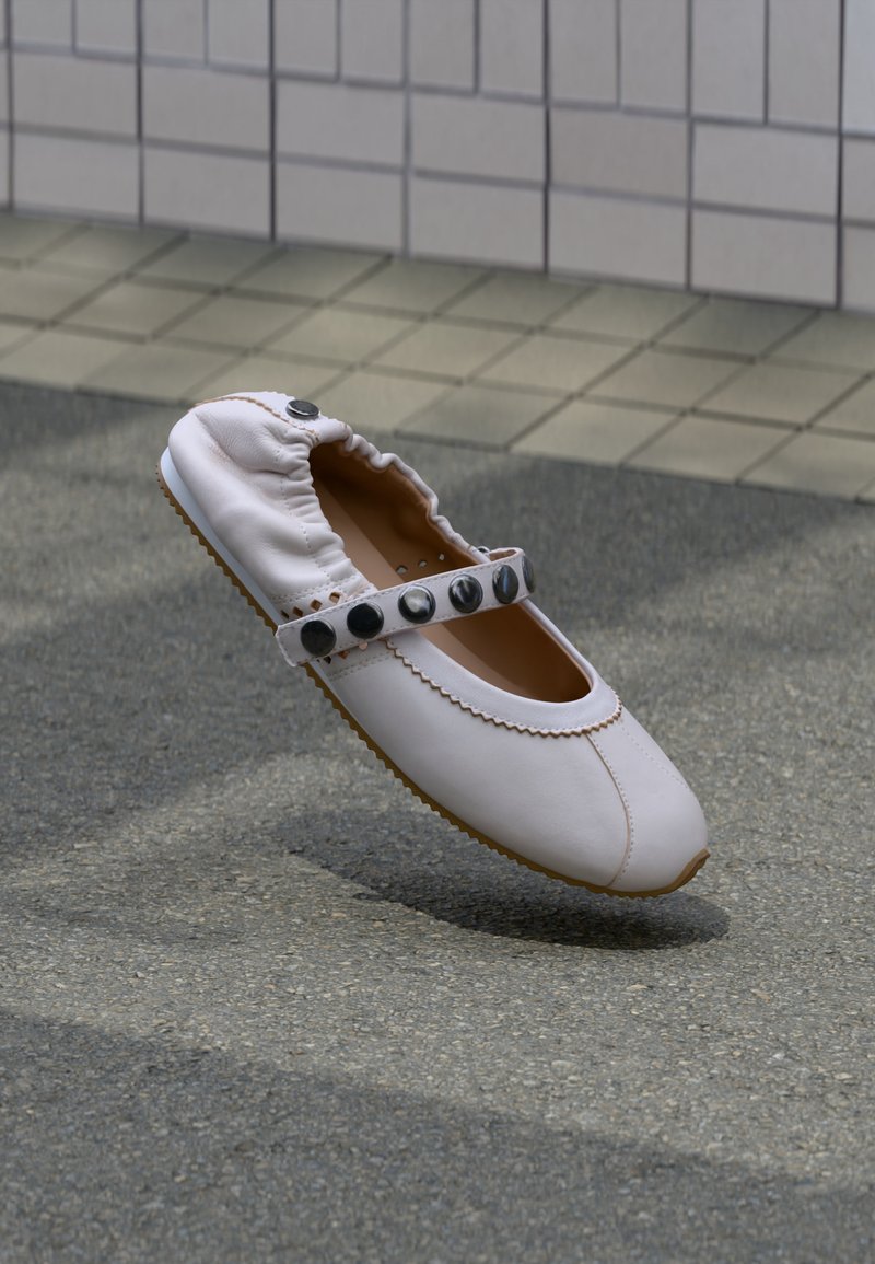 Λευκό δερμάτινο παπούτσι με επίπεδη σόλα, ελαστικό πίσω μέρος και λουράκι διακοσμημένο με μεγάλα μαύρα κουμπιά, πάνω σε γκρίζο άσφαλτο με φόντο τοίχο με πλακάκια.
