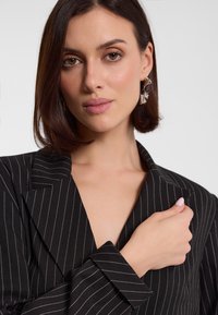 Czarny żakiet w prążki z klapami i długimi rękawami. Model trzyma rękę na kołnierzu, prezentując elegancki, dopasowany krój oraz subtelną teksturę.