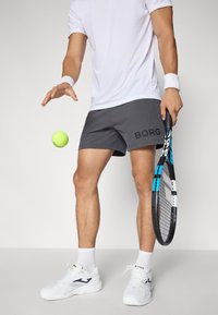 Hallid hallid sportlikud lühikesed püksid "BORG" logo ja valge lühikeste varrukatega särk, hoides tennisereketit ja rohelist tennisepalli. Valged tossud mustade detailidega.