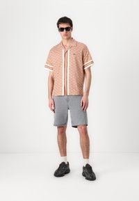 Kortermet skjorte i oransje og beige geometrisk mønster med mørke striper, parret med grå denimshorts og svarte joggesko. Sorte solbriller.