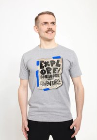 Šedé tričko s krátkým rukávem s grafikou zmuchlaného papíru a černým textem "PROZKOUMEJ! JEŠTĚ VÍC DOBRODRUŽSTVÍ", přilepeným modrou páskou.