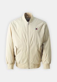 Lys beige bomberjakke med glidelås, ribbede mansjetter og avslutning. Har en liten logo-lapp i rødt, hvitt og blått på brystet.