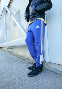 Niebieskie spodnie dresowe Adidas z białymi paskami po bokach, w zestawieniu z czarnymi trampkami, noszone z czarną skórzaną kurtką i żółtą koszulą.