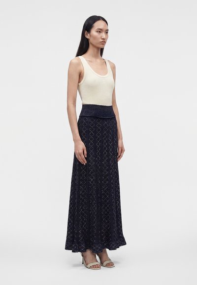 Femme debout en débardeur crème sans manches, longue jupe bleu marine à motifs et sandales à talons claires sur un fond uni clair.