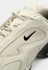 Κρέμα χρώματος αθλητικά παπούτσια Nike με υφασμάτινες επιφάνειες, μαύρο λογότυπο swoosh και λευκές κορδόνες σε λευκό φόντο.
