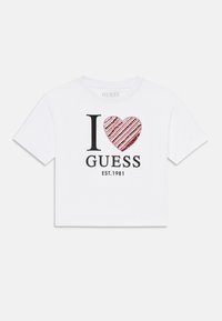 Valge lühikeste varrukatega särk mustade "I" ja "GUESS" tekstidega; punane südamemuster, mis on täidetud sektsiooniga ja triipudega; asutamise aasta 1981.