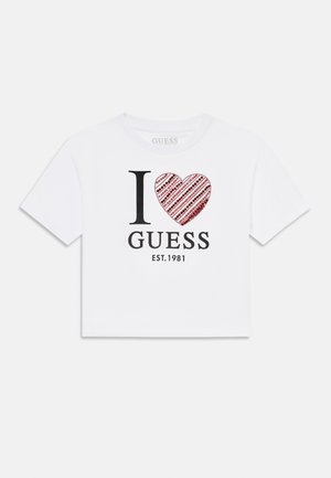 T-shirt blanc court avec le texte noir "I" et "GUESS" ; design de cœur rouge rempli de paillettes et de rayures ; année d'établissement 1981.