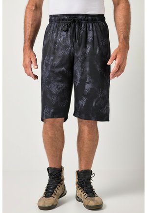 Mand iført sorte mønstrede træningsshorts og lysebrune høje sneakers står foran en ensfarvet baggrund.