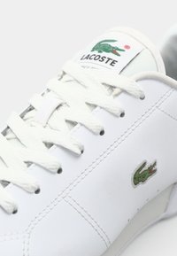 Baskets blanches Lacoste avec logo crocodile vert sur le côté et la languette, lacets blancs et détails cousus sur la tige en cuir lisse.