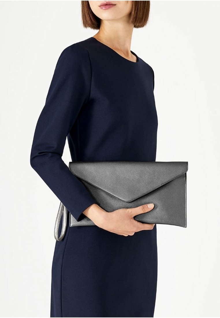 Silver leather clutch with an envelope design, smooth texture, and a thin strap. Held by a person in a fitted navy dress.