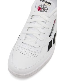 Lähedalt vaadatuna valge Reebok Classic spordijalats, millel on perforatsioonidega varvas, valged sidemed, must küljektähis ning Reebok logo punase ja musta detailiga.