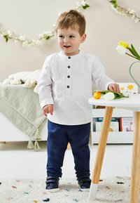 Witte shirt met decoratieve borduursels en zwarte knopen, gecombineerd met een marineblauwe broek en donkere schoenen. Kind staat naast een tafel.