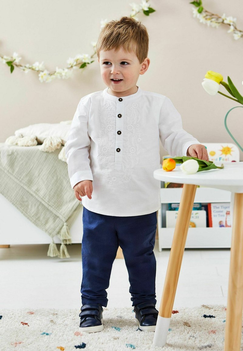 Witte shirt met decoratieve borduursels en zwarte knopen, gecombineerd met een marineblauwe broek en donkere schoenen. Kind staat naast een tafel.
