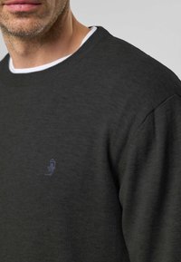 Tummanvihreä neulottu pusero pyöreällä kaula-aukon reunuksella, jossa pieni, yksityiskohtainen kirjailtu logo rinnassa. Pehmeä pinta ja rento istuvuus.