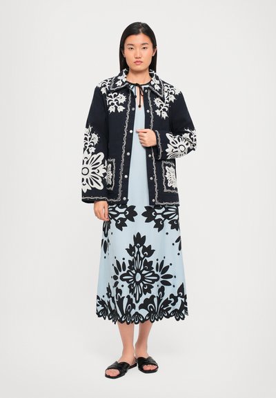 Schwarz-weiße floral gestickte Jacke über einem hellblauen Kleid mit schwarzen Blumenmustern, kombiniert mit schwarzen Sandalen.