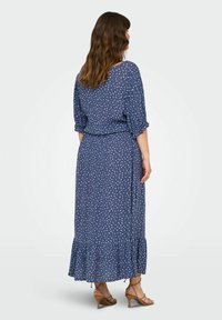 Abito lungo di colore navy con pois bianchi. Tessuto fluido, maniche corte, vita arricciata e orlo con volants. Tacchi a strappo marroni visibili.