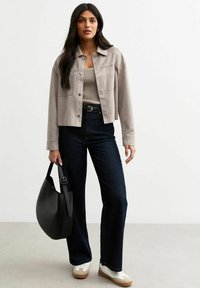 Grijze suède cropped jack over een beige ribbeltop, gecombineerd met donkerblauwe flared jeans en witte sneakers. Houdt een zwarte hobo tas vast.