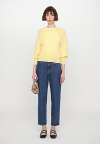 Gelber gestrickter Pullover mit Raglanärmeln, kombiniert mit blauen Jeans im geraden Schnitt. Hält eine kleine Musterhandtasche und trägt schwarze Schuhe.