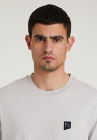 Světle šedé tričko s kulatým výstřihem vyrobené z jemné látky, s malou černou nášivkou na levé části hrudi a jednoduchým designem.