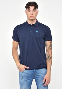 Námořnická modrá polo tričko vyrobené z bavlny s hladkou texturou, se zapínáním na tři knoflíky a malým modrým logem na hrudi.