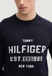 Granatowy sweter z dzianiny z białym napisem: "TOMMY HILFIGER EST. 1985 NEW YORK." Okrągły dekolt i ściągacze przy rękawach.
