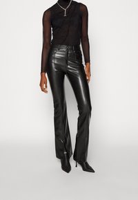 Svarta åtsittande läderbyxor med hög midja och utsvängda ben, kombinerat med en genomskinlig svart långärmad topp med volangdetaljer.