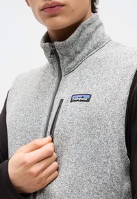 Άτομο που φοράει γκρι φλις γιλέκο Patagonia με μαύρα μανίκια, κρατώντας και ξεκουμπώνοντας το φερμουάρ στην τσέπη στο στήθος.