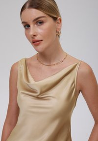 Camiseta de satén en color crema con escote drapeado y tirantes finos. Llevada con un collar choker en tono dorado y pendientes de aro, mostrando una textura suave.