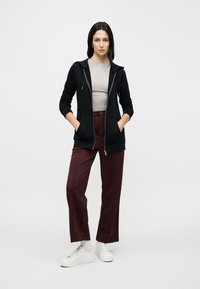 Black zip-up hoodie with a drawstring, fitted gray shirt, maroon wide-leg trousers, and white sneakers on a plain background.