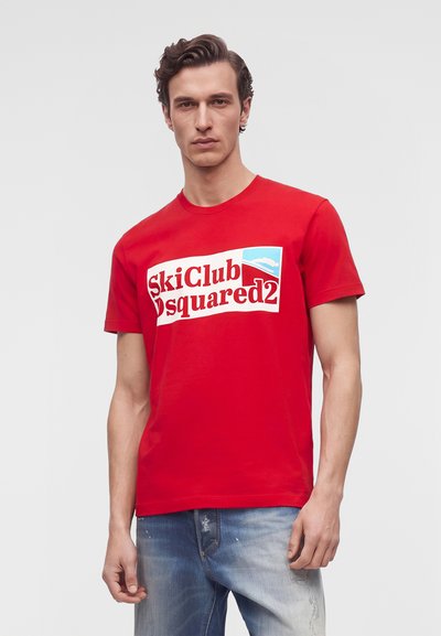 Мъж позира с червена тениска Ski Club Dsquared2 и сини дънки на светъл едноцветен фон.