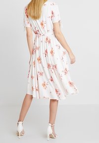 Blommig midiklänning med korta ärmar, vit bakgrund, rosa och orange blommor, insvängd midja, flödande kjol, matchad med vita klacksandaler.