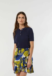 Marineblauwe polo met korte mouwen en ribbels, voorzien van drie knopen, gecombineerd met een gele en blauwe rok met ruffles die voorzien is van bloemen- en golfpatronen.