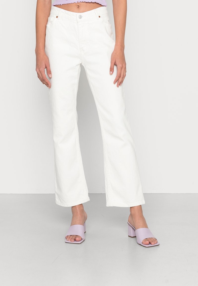 Personne portant un jean blanc taille haute à jambes larges et des sandales à talons ouvertes violet clair, debout devant un fond uni.