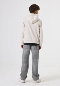 Sudadera beige clara con capucha, llevada sobre una camiseta negra, combinada con unos pantalones vaqueros gris holgados. Zapatillas blancas visibles. Diseño simple, ajuste casual.