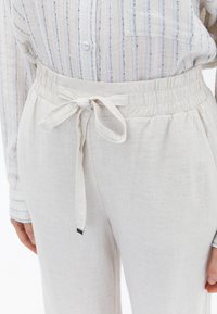 Lichte, crèmekleurige broek van een textuur materiaal. Heeft een elastische tailleband met een strik en een subtiele glans.