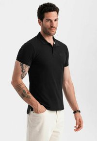 Svart polo skjorta med krage och korta ärmar, som har en subtil logotyp på bröstet och en strukturerad tyg. Bärs tillsammans med beige byxor.