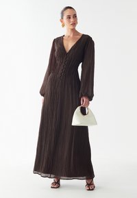 Rjava, teksturirana maksi obleka z globokim V-izrezom, dolgimi rokavi in gumbom po sprednji strani; v paru z majhno belo torbico.