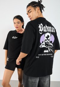 Black oversized t-shirt with Batman graphic on back, featuring purple accents and white text. Soft cotton fabric with relaxed fit.