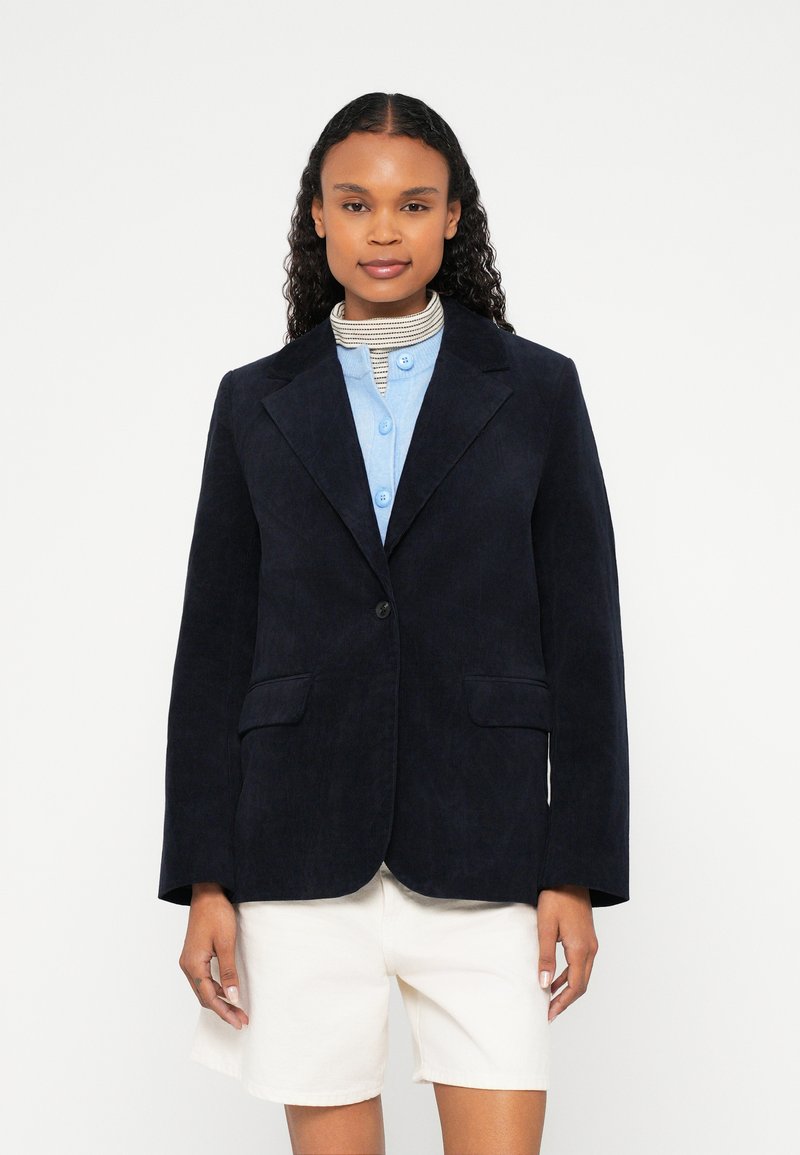 Mornarski džinsasti blejzer s jednim gumbom, reverima s urezom, dva prednja džepa, nošen preko svijetloplave košulje s kopčanjem i bijelih kratkih hlača.