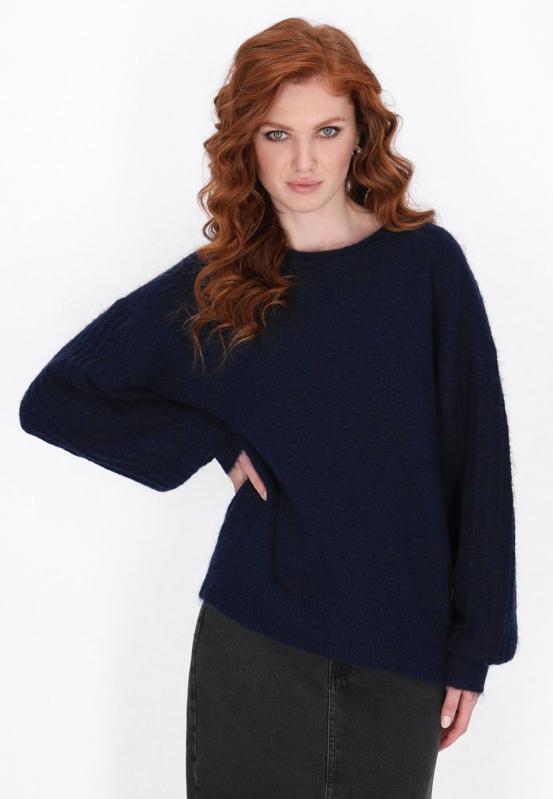 Marineblaues Strickpullover mit lockerer Passform, weitem Bootsausschnitt, gerippten Ärmeln und einer weichen, strukturierten Oberfläche. Kombiniert mit einem schwarzen Rock.