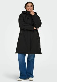 Svart vadderad jacka med huva, dragkedjefickor och en avslappnad passform. Kombinerad med blå utsvängda jeans och vita skor. Slät textur, midjelång design.