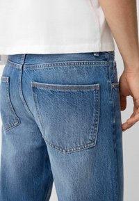 Jeans de mezclilla azul con una textura ligeramente desvanecida, dos bolsillos traseros, detalles de costura y un corte recto. Llevados con una camisa blanca.